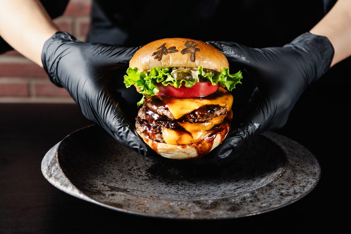 Wagyu burger with cheese, tomato and lettuce on a bun branded 和牛, held in gloved hands